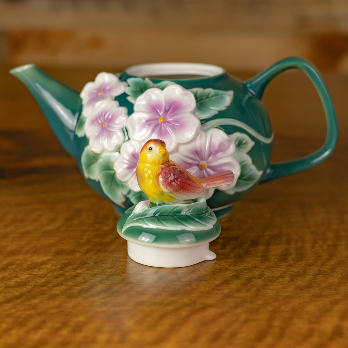 Teal teapot with raised pink flowers and a yellow bird ornament on a wooden surface.