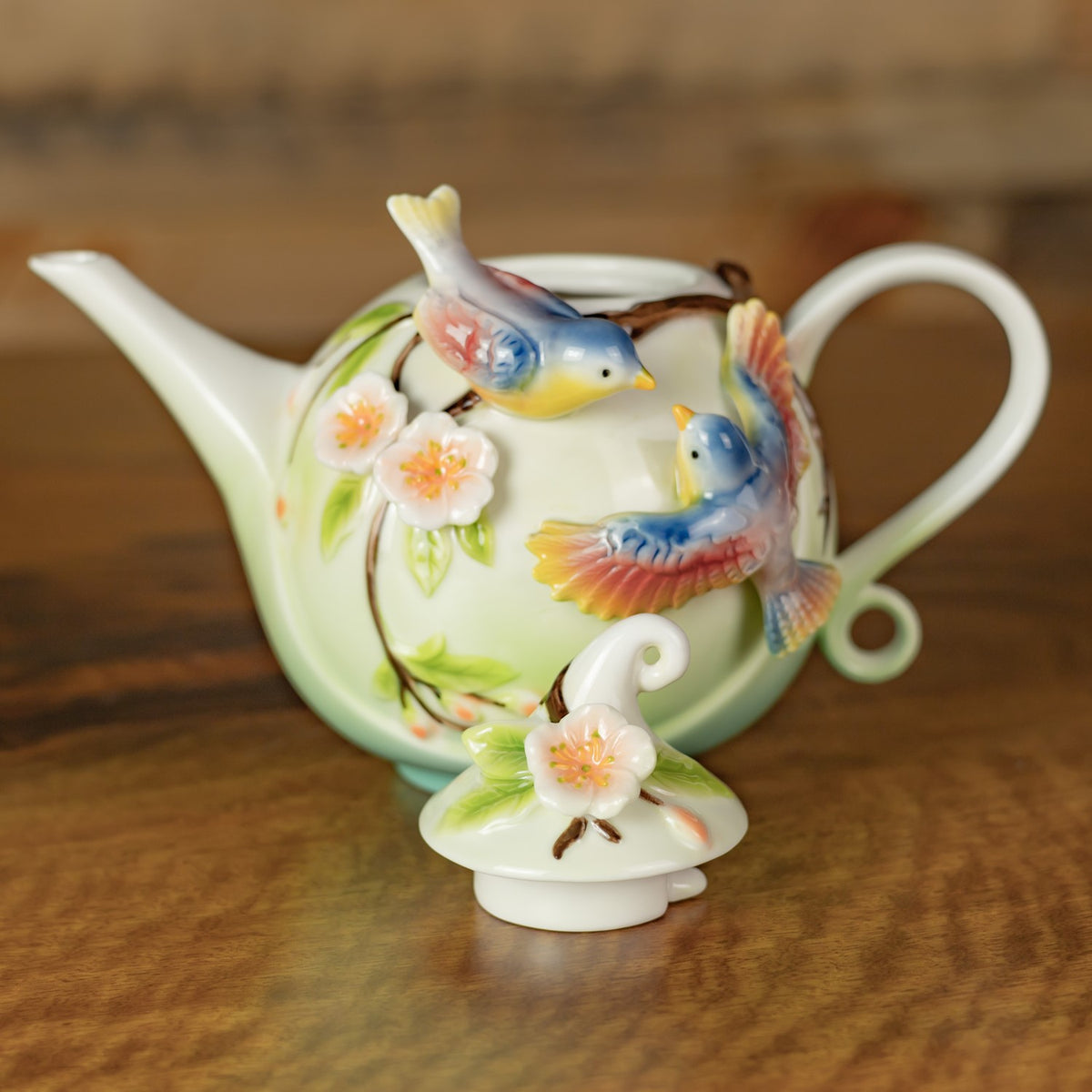 Decorative teapot with colorful birds and blossoms, shown with its flower-decorated lid resting in front.