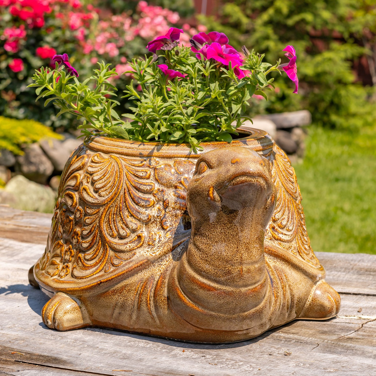 Ornately patterned turtle planter filled with bright pink flowers on a wooden surface in a garden.