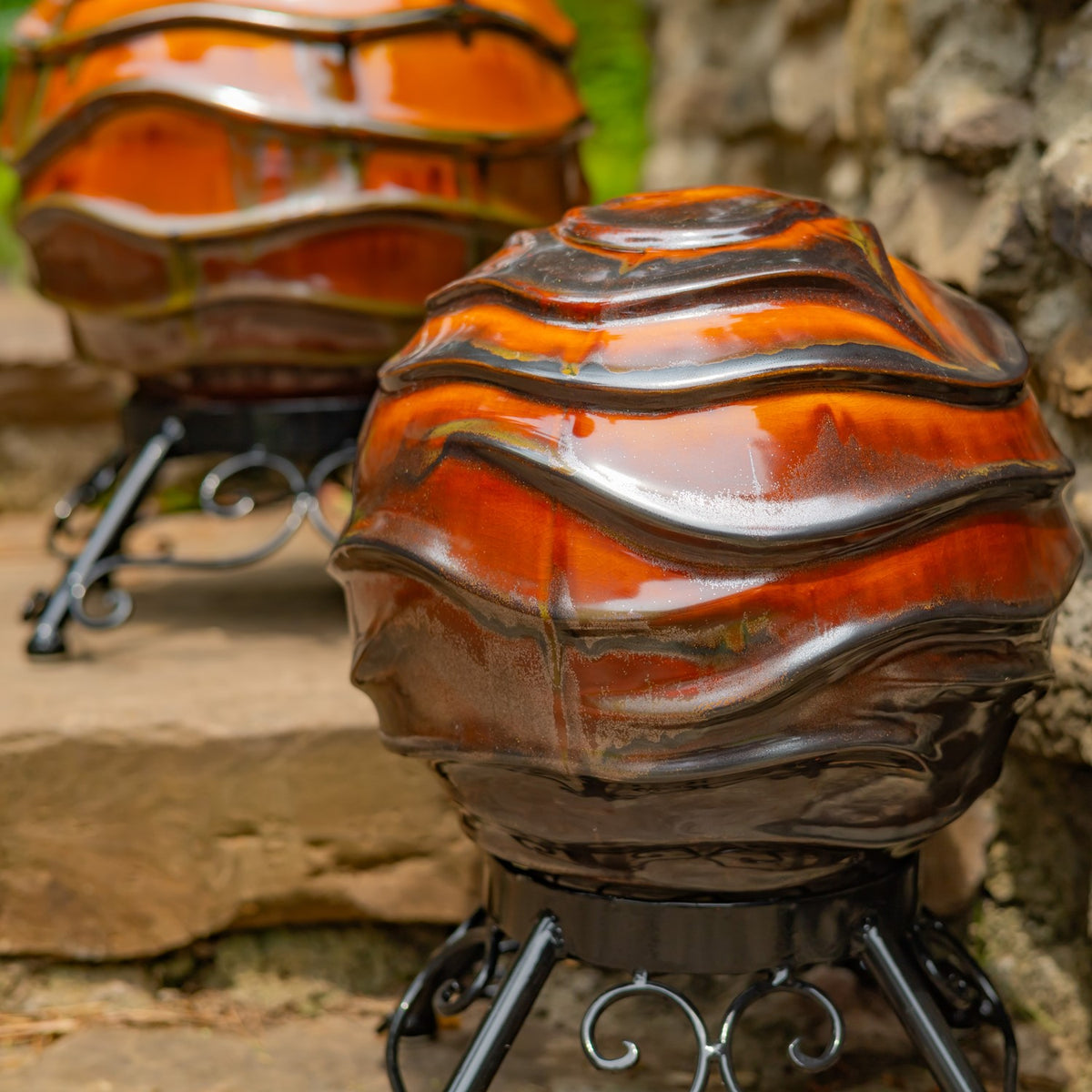 Two glossy orange-and-brown wavy globes on black scrollwork stands beside a stone wall.