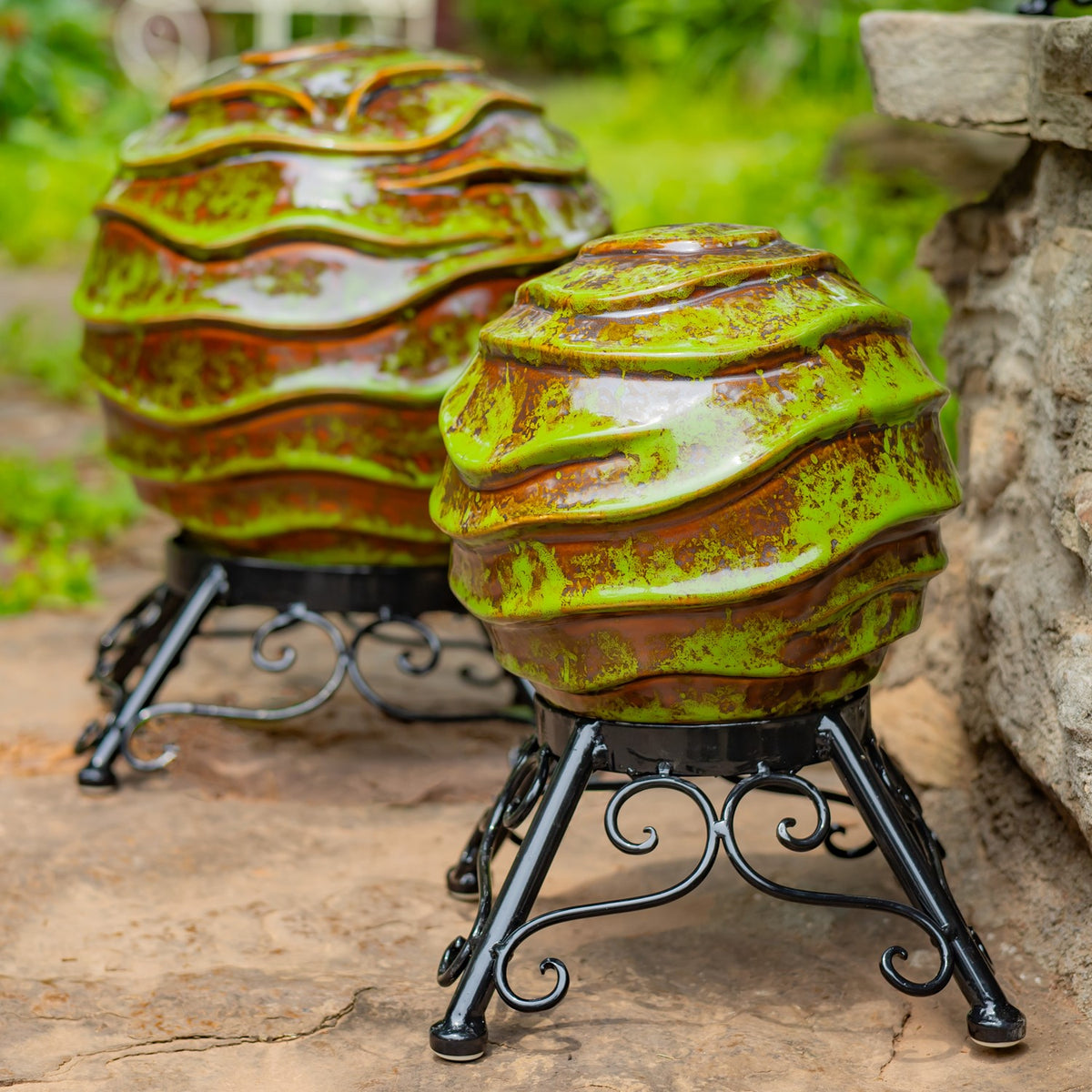 Two glossy green and brown patterned gazing globes sit on black scrollwork metal stands in a garden.