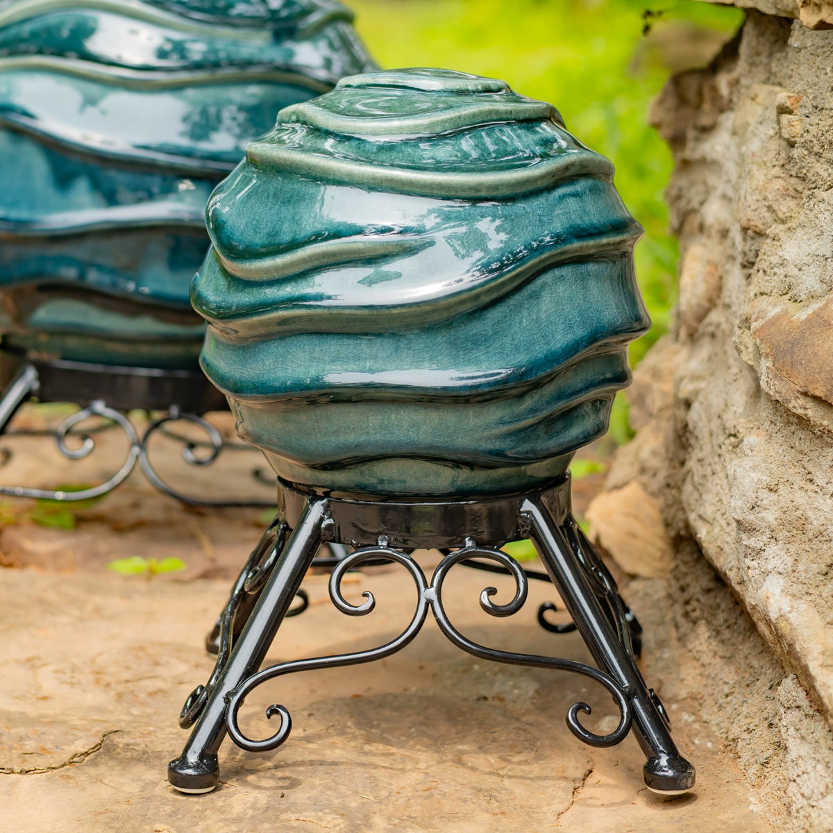 A teal glazed globe with raised bands sits on an ornate black metal stand outdoors.