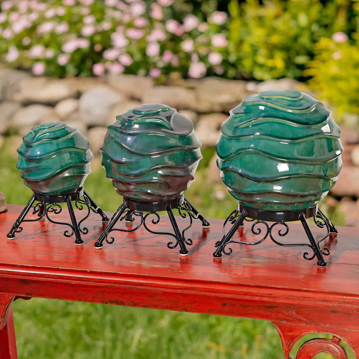Three glossy teal gazing globes in different sizes sit on ornate black metal stands on a red bench.