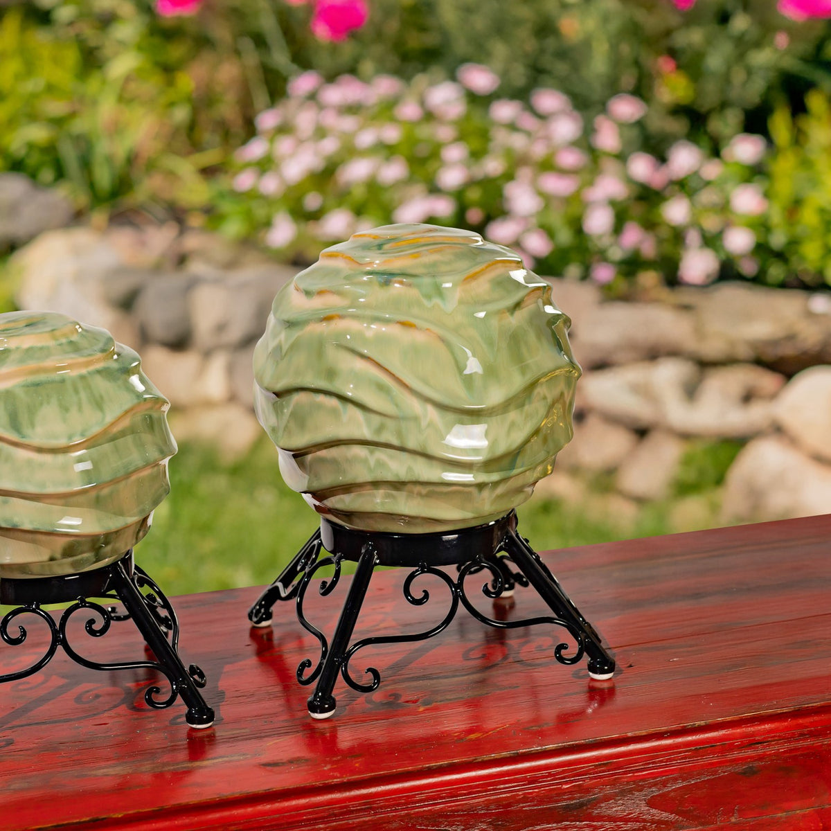 Two pale green, wavy-patterned gazing globes on black metal stands sit on a red wooden table in a garden.