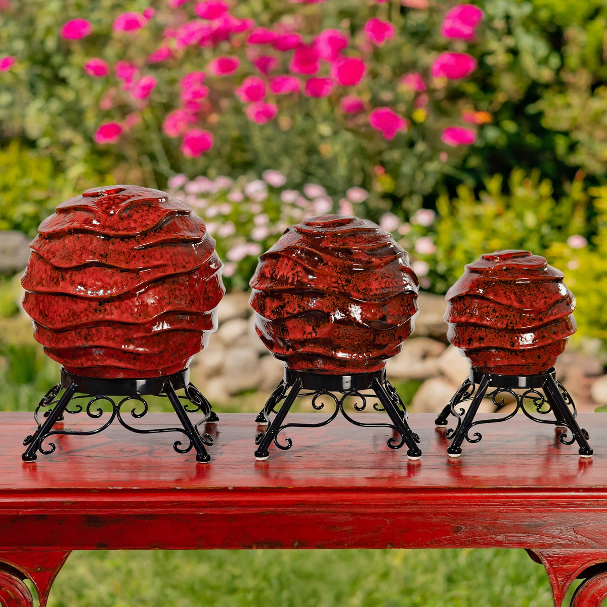 Three red textured gazing globes on black metal stands displayed on a garden table.