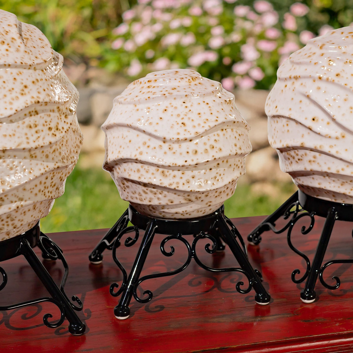 Three cream speckled porcelain globes on black scrollwork stands, arranged on a red table in a garden.