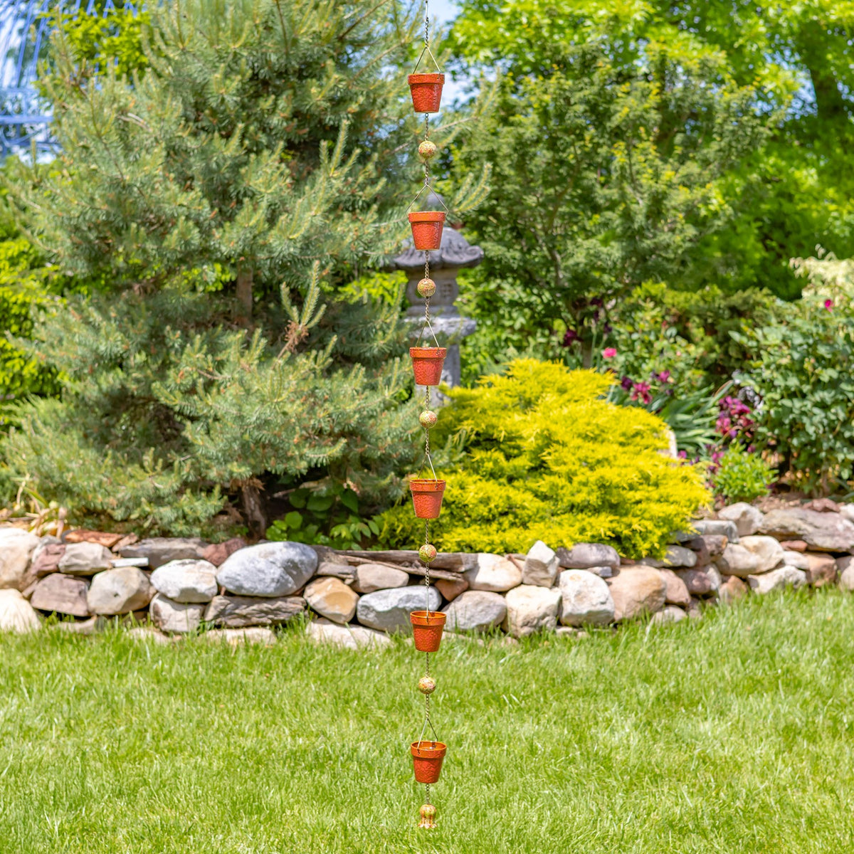 Red bucket rain chain hangs in a garden above grass, with shrubs and a low stone border behind it.