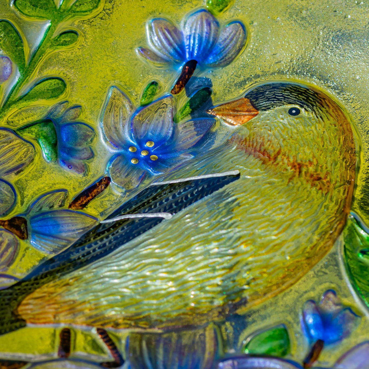 Close-up of a goldfinch and blue flowers embossed in a colorful glass bowl.