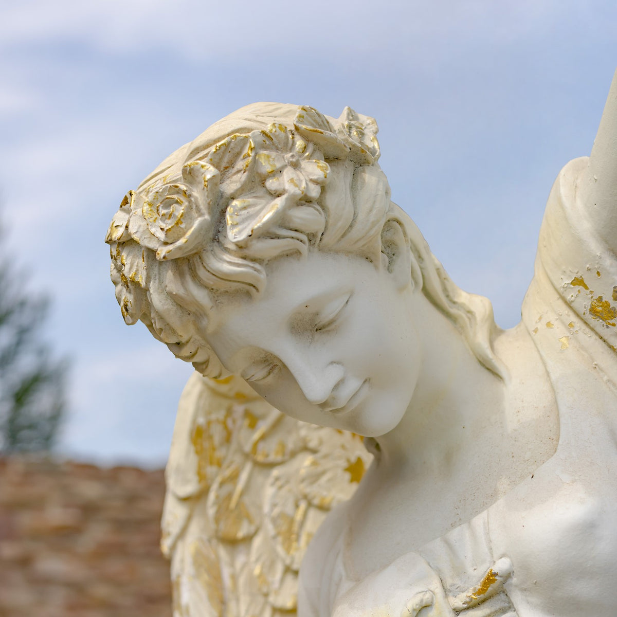Close-up of the angel statue’s bowed face, carved curls, and gold-flecked wing against the sky.