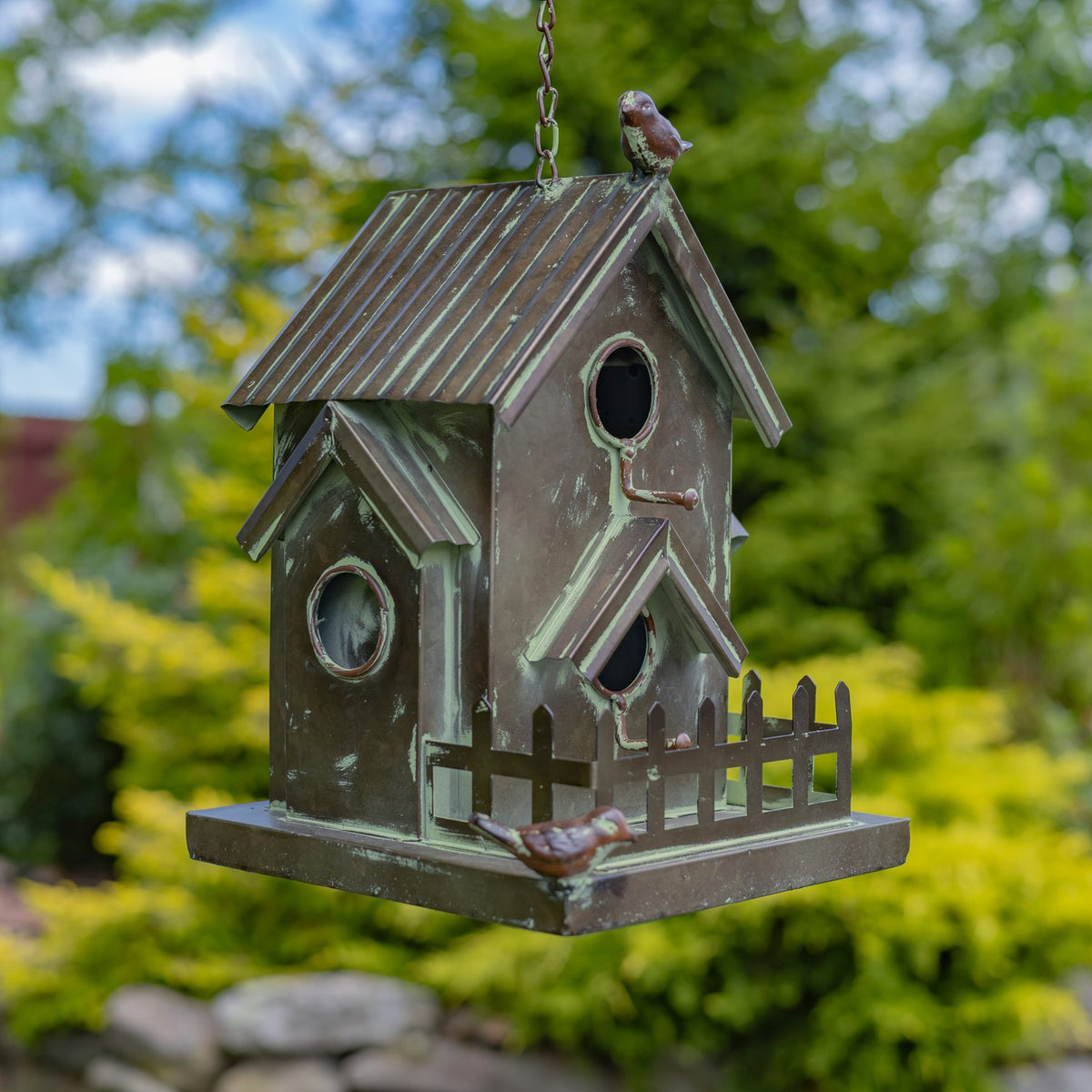 A weathered metal birdhouse hangs outdoors, with peaked roofs, round openings, a small fence, and bird ornaments.