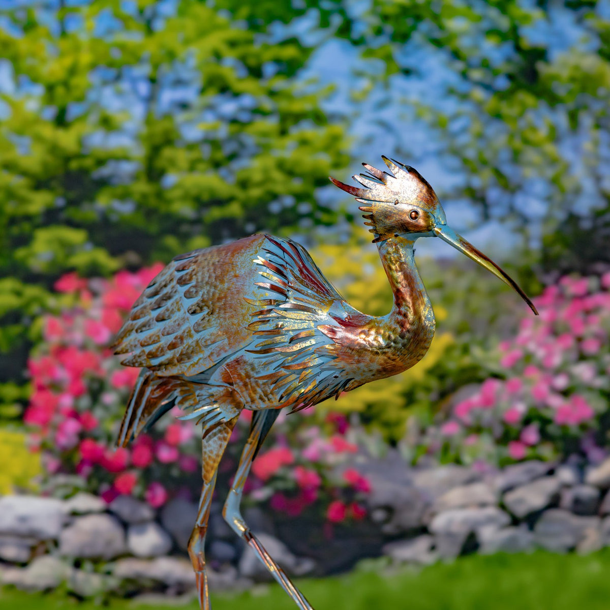 Metal heron figurine with textured wings standing among flowers and rocks in a garden.