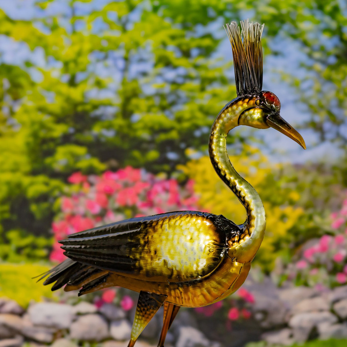 Metallic heron figurine with a curved neck and long beak, standing against a flower-filled garden backdrop.