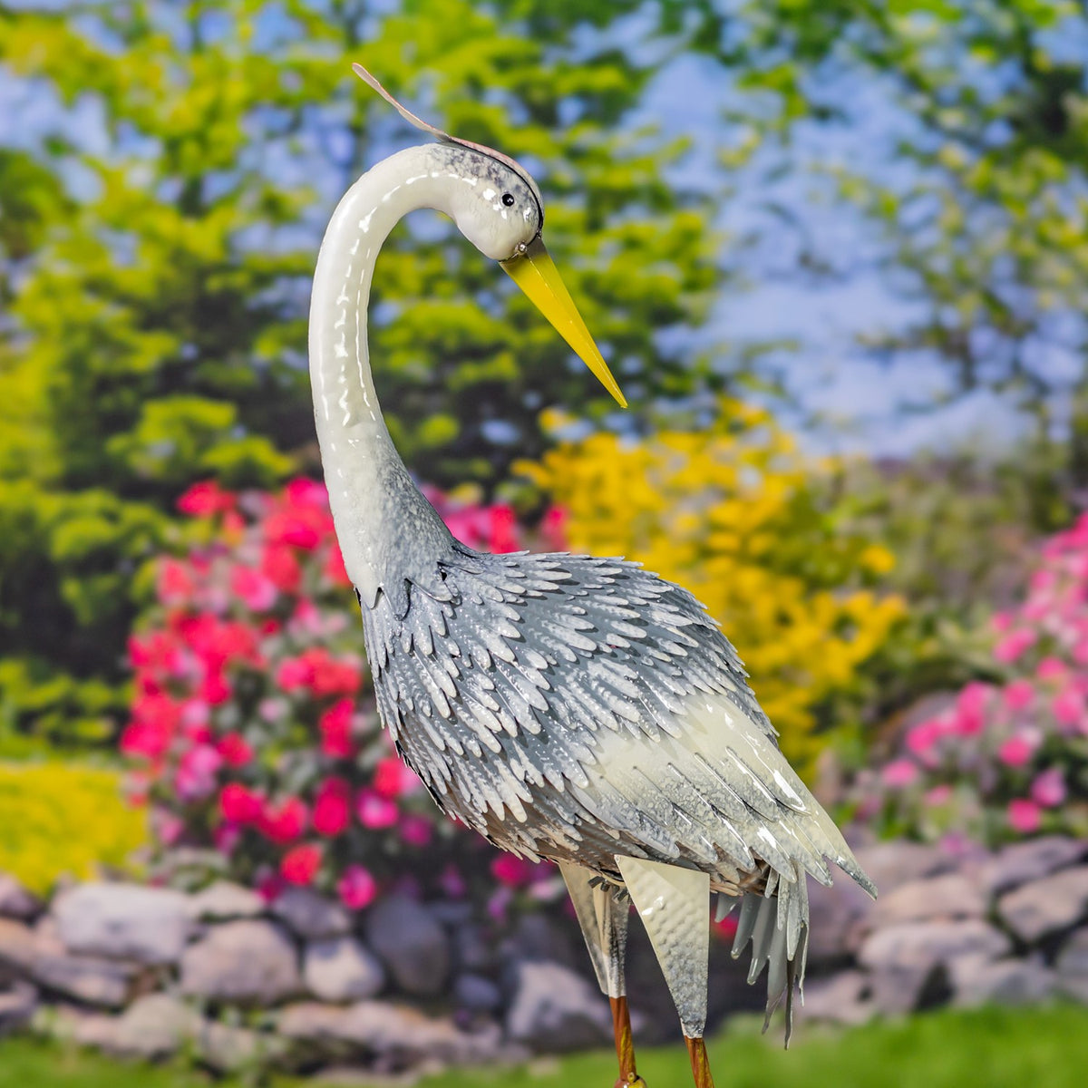 A metal heron figurine with a curved white neck, yellow beak, and layered gray feathers stands among garden flowers.