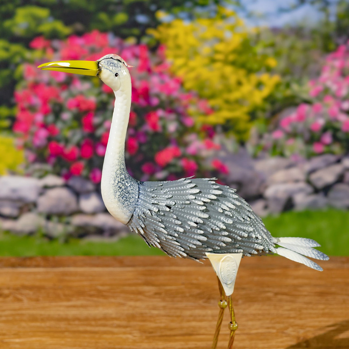 Metal heron figurine standing upright with a long yellow beak and layered gray wings against a garden backdrop.