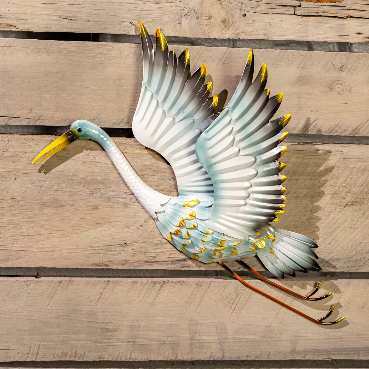 Metal crane wall decoration with raised wings, pale blue and white feathers, yellow accents, and long legs on a wood wall.