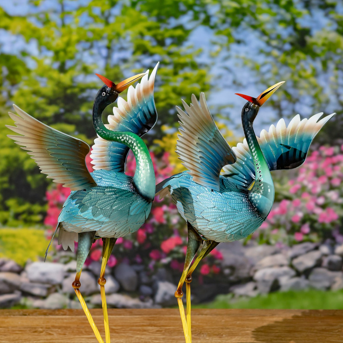 Two blue heron figurines stand with wings spread on a wooden surface in a flower-filled garden.