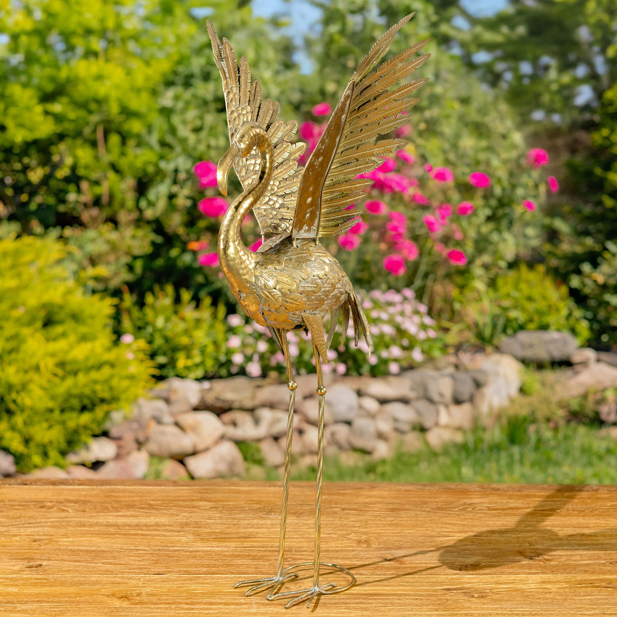 Gold flamingo figurine with raised wings standing on a wooden surface in a flower garden.