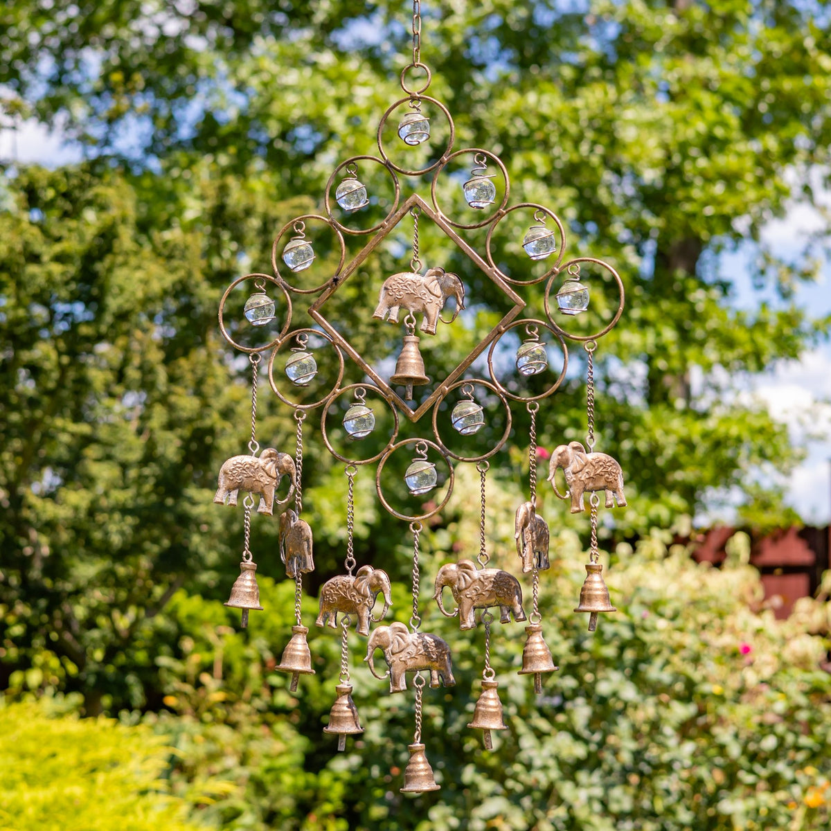 Ornate bronze-tone wind chime with elephant figures and clear hanging accents against a leafy garden.