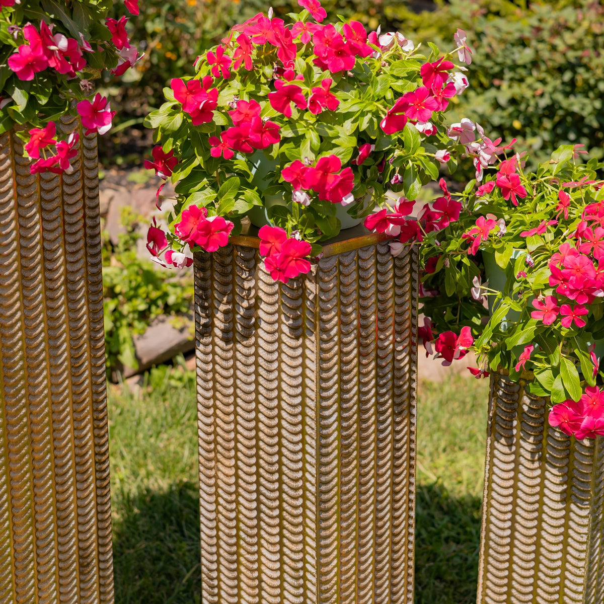 Three textured gold-toned pedestal pillars hold lush planters of bright pink flowers in a garden.