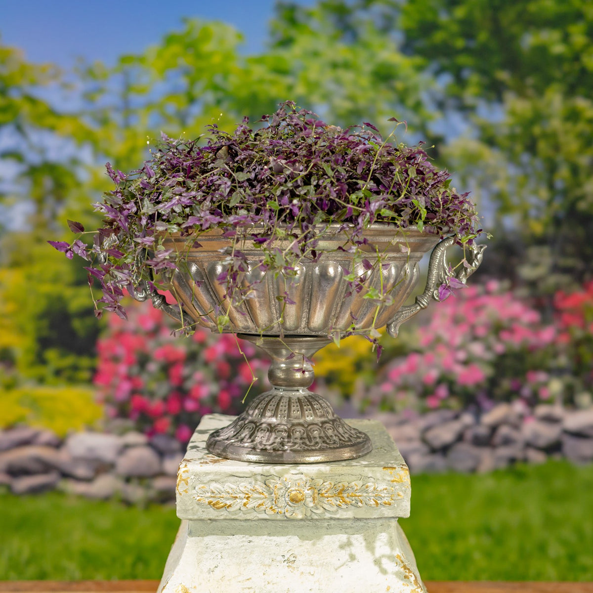 Ornate silver urn filled with purple foliage on a pedestal in a garden setting.