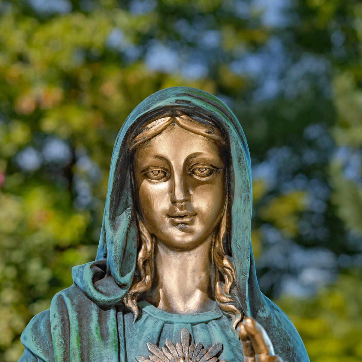 Close view of Mary's face and blue-green veil, with part of the Sacred Heart visible against a garden backdrop.