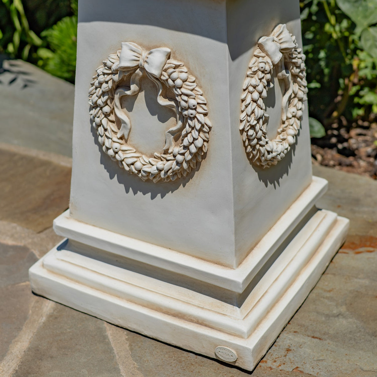 Close view of the pale pedestal base, showing raised bow-tied wreaths on two sides and stepped trim.