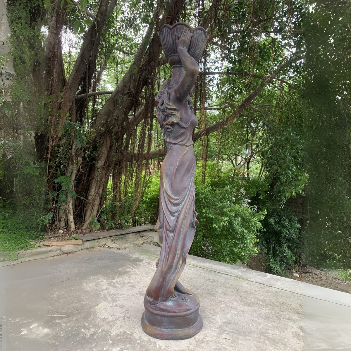 A draped female statue stands on a round base, raising a flower pot overhead beside a leafy garden.