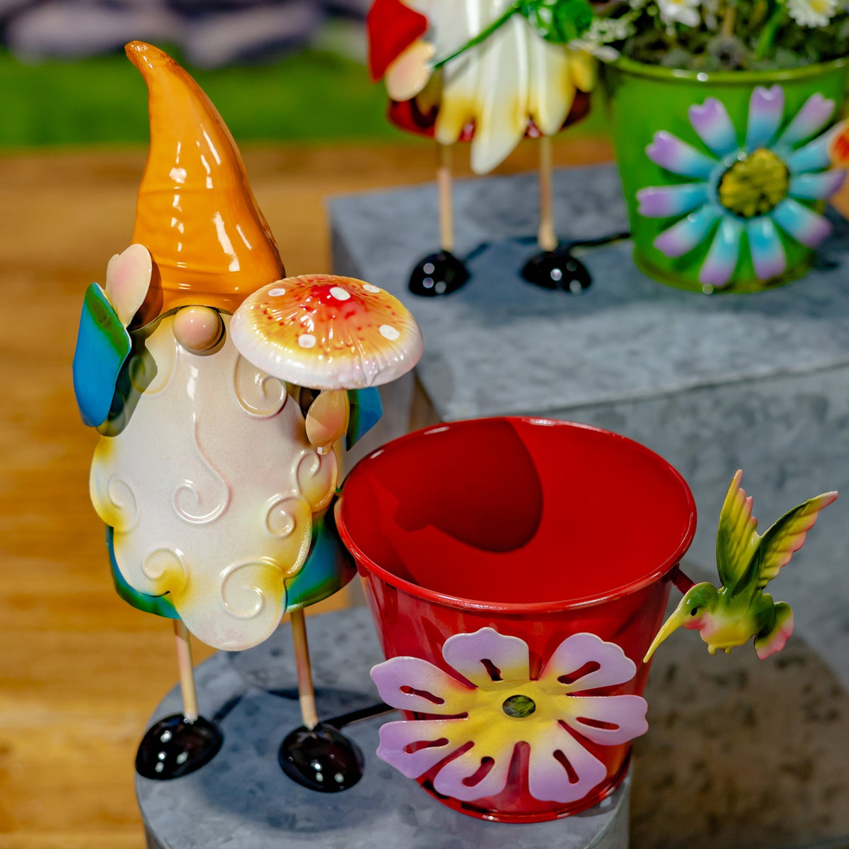 A white-bearded gnome with an orange hat stands beside a red flower pot decorated with a pink flower.