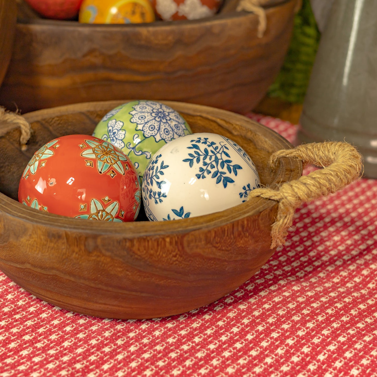 A round wooden tray with rope handles holds three patterned decorative eggs on a red checked cloth.