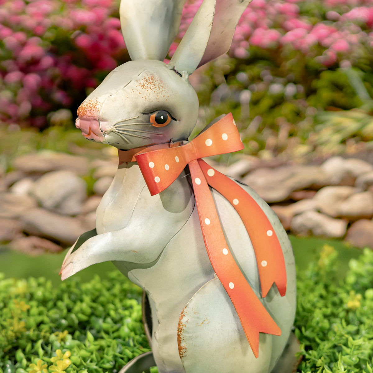 Close view of a pale metal rabbit figurine wearing an orange polka-dot ribbon among garden plants.
