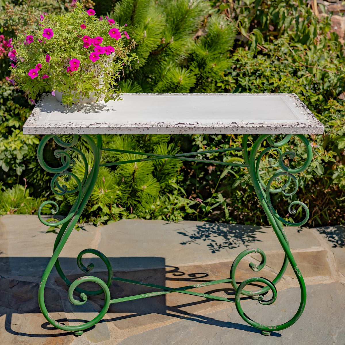 Rectangular white-topped table with ornate green iron legs, shown outdoors beside flowering plants.