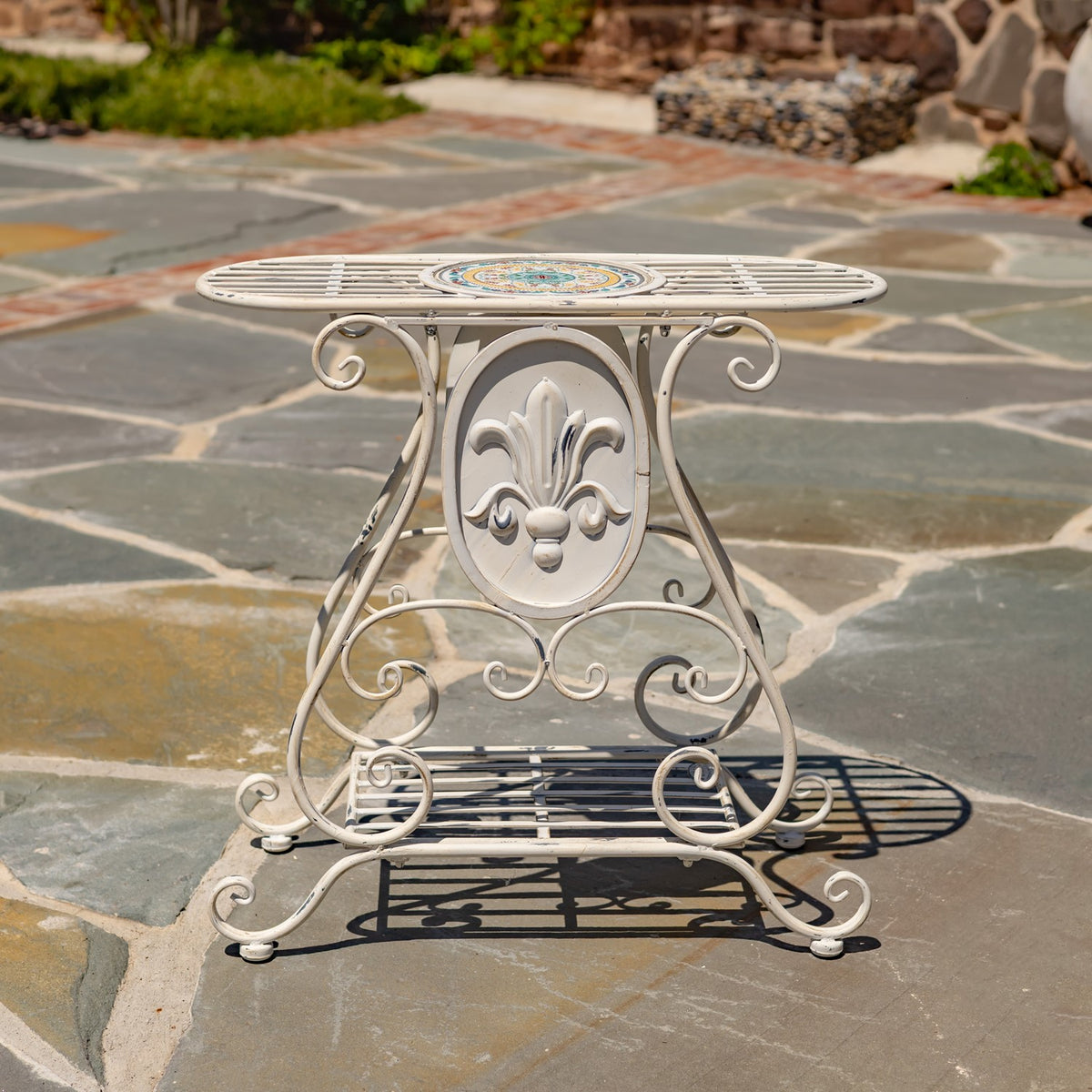 Cream metal plant stand with a colorful mosaic tabletop and ornate scrollwork on a stone patio.