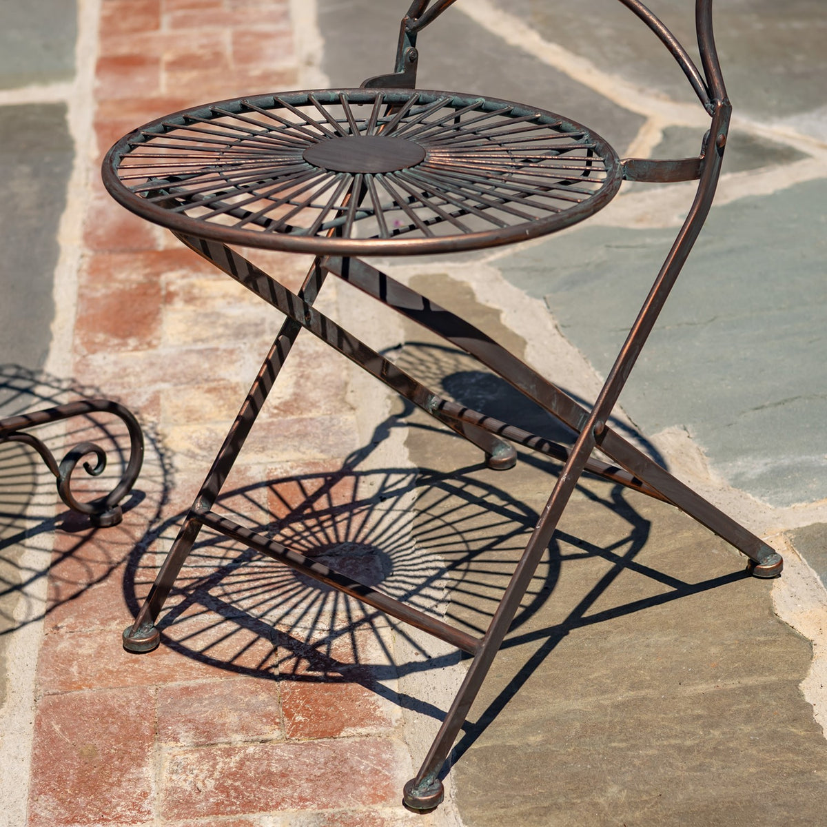 A dark metal wire-frame bistro chair stands on stone paving, casting a spoke-like shadow.