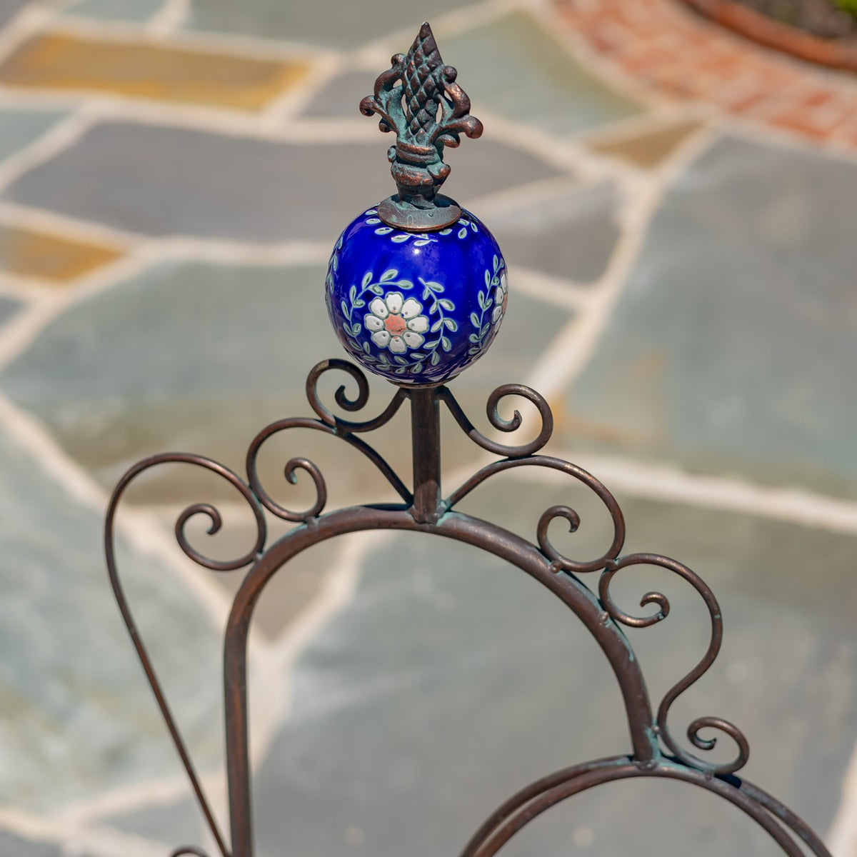 Close-up of an ornate bronze scrollwork frame topped with a blue floral-patterned ceramic orb and finial.