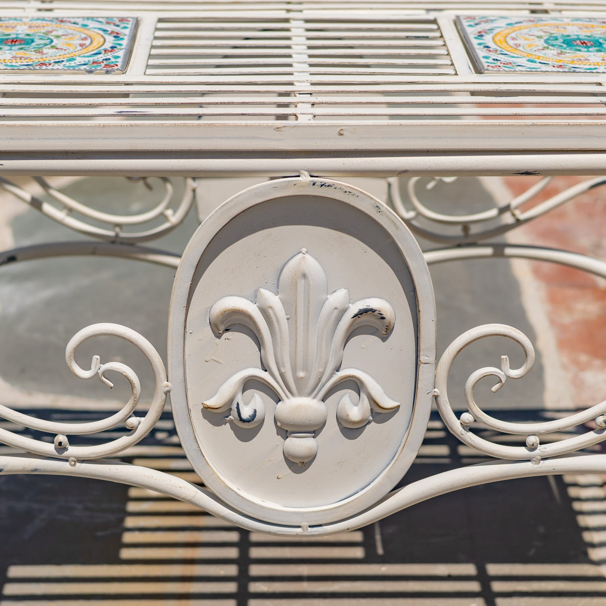 Close-up of an ornate white metal table apron with a raised fleur-de-lis medallion and curling scrollwork.