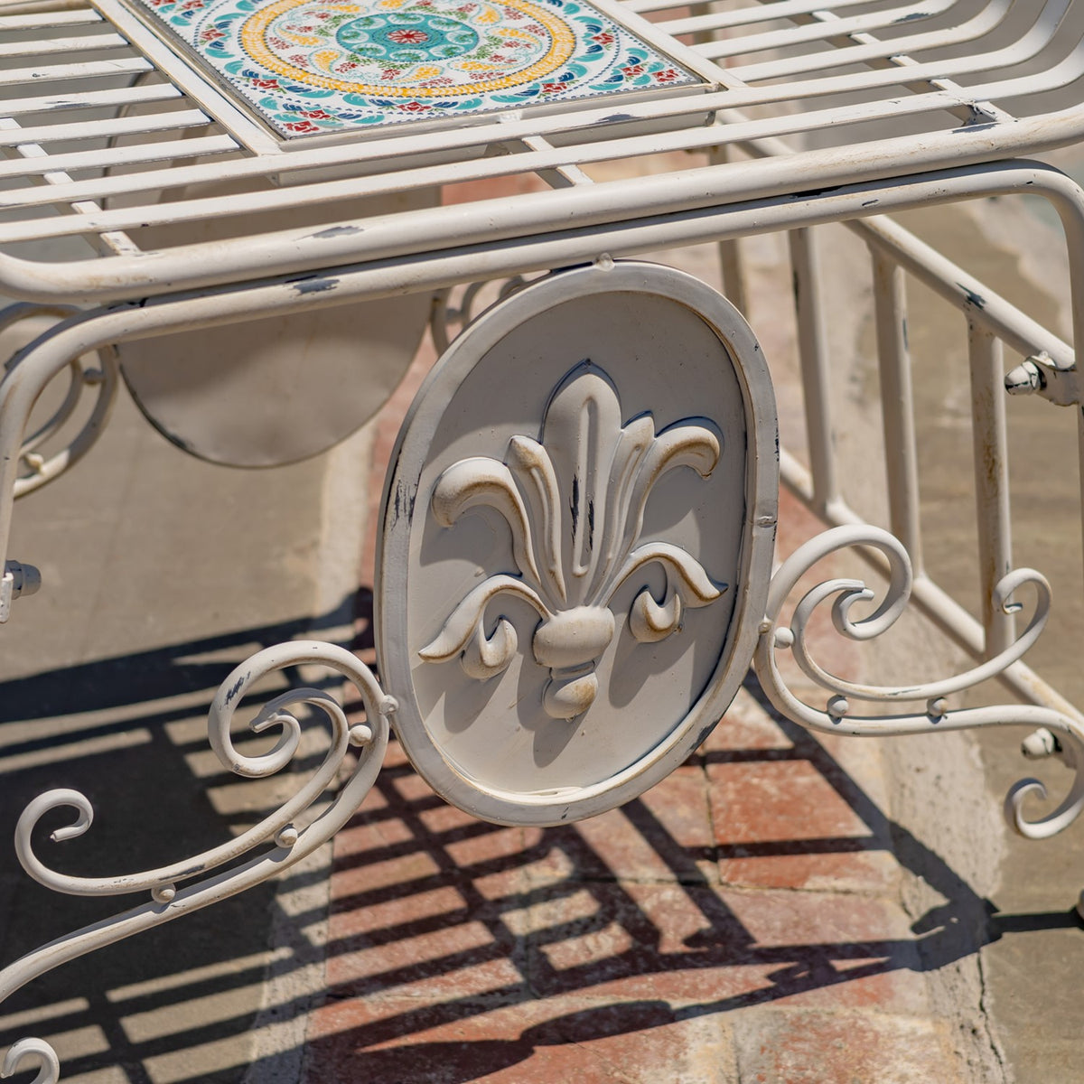 Close-up of an ivory metal chair with a raised fleur-de-lis medallion and colorful mosaic seat.