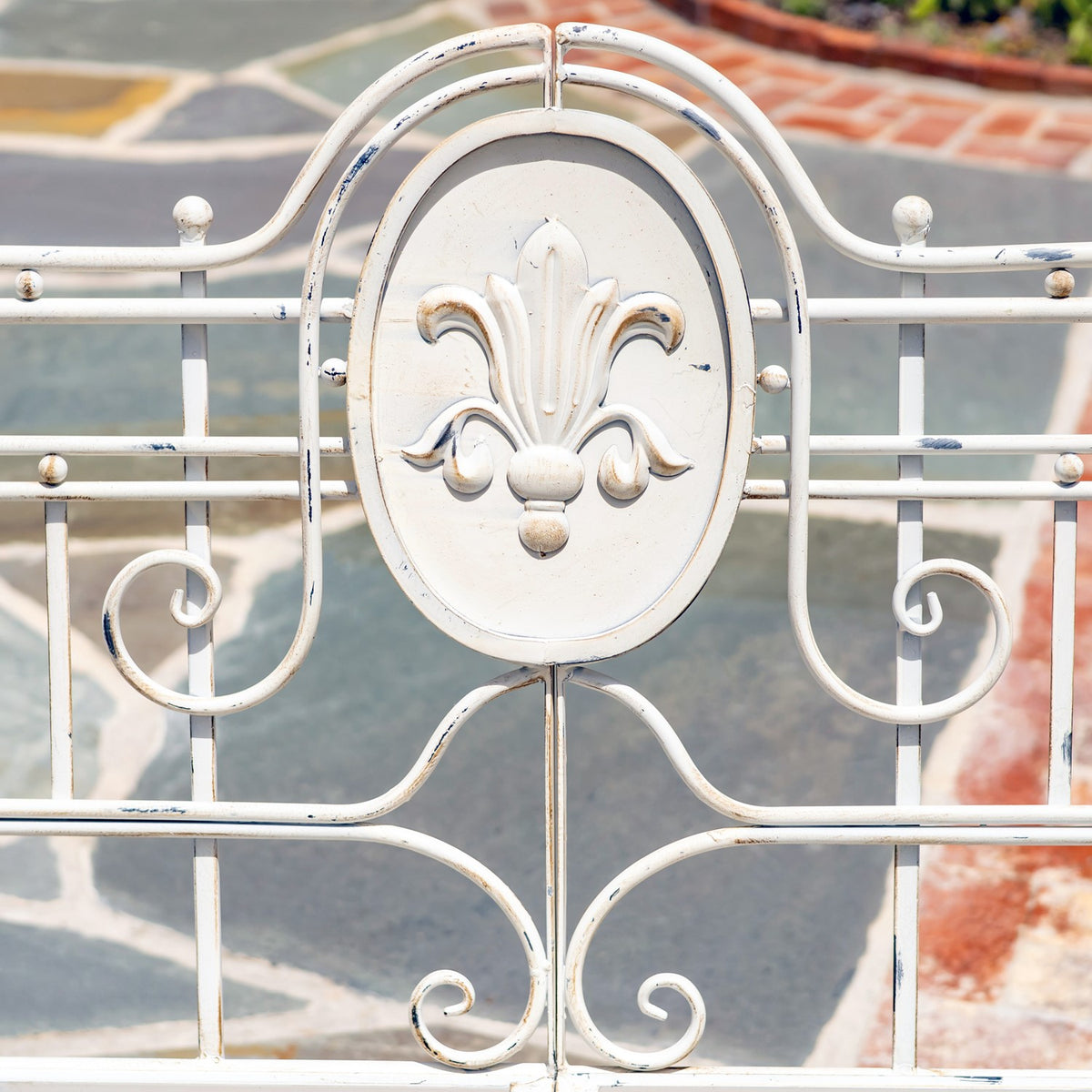 Close-up of the bench’s white metal backrest with a raised fleur-de-lis medallion and curling scrollwork.