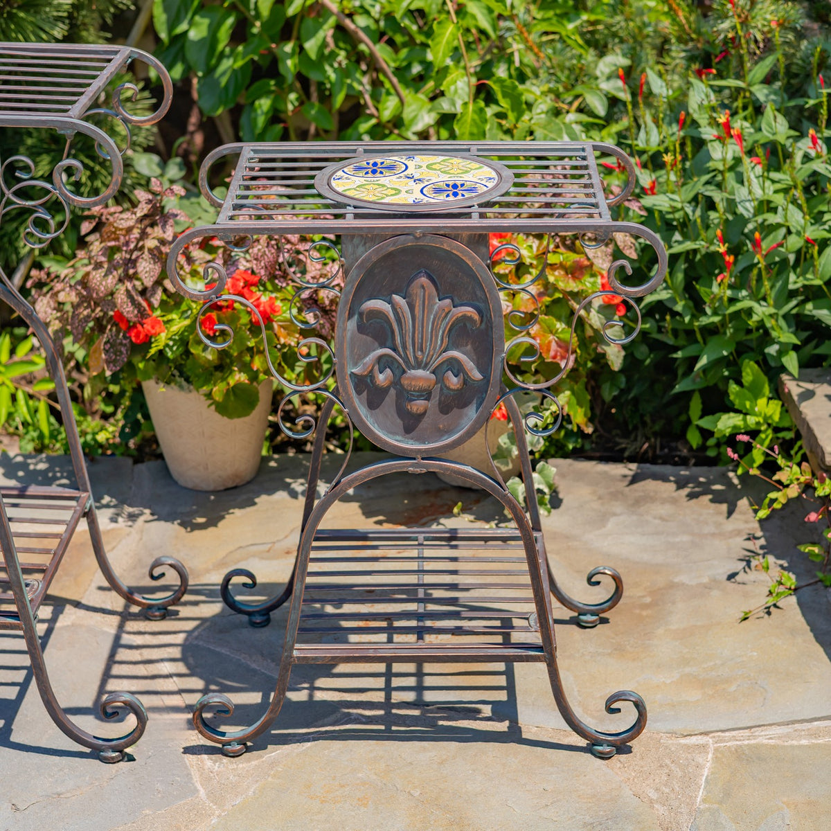 Ornate metal plant stand with a colorful mosaic top and fleur-de-lis panel, shown among garden plants.