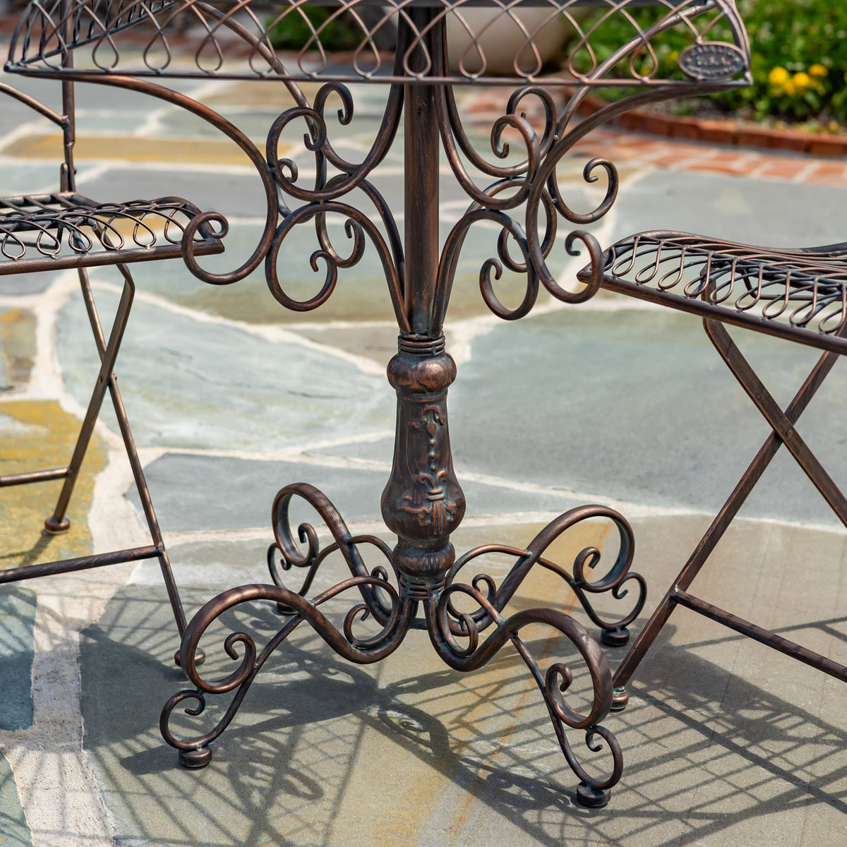 Close view of a dark metal bistro table base with curled scrollwork between two matching chairs on a tiled patio.