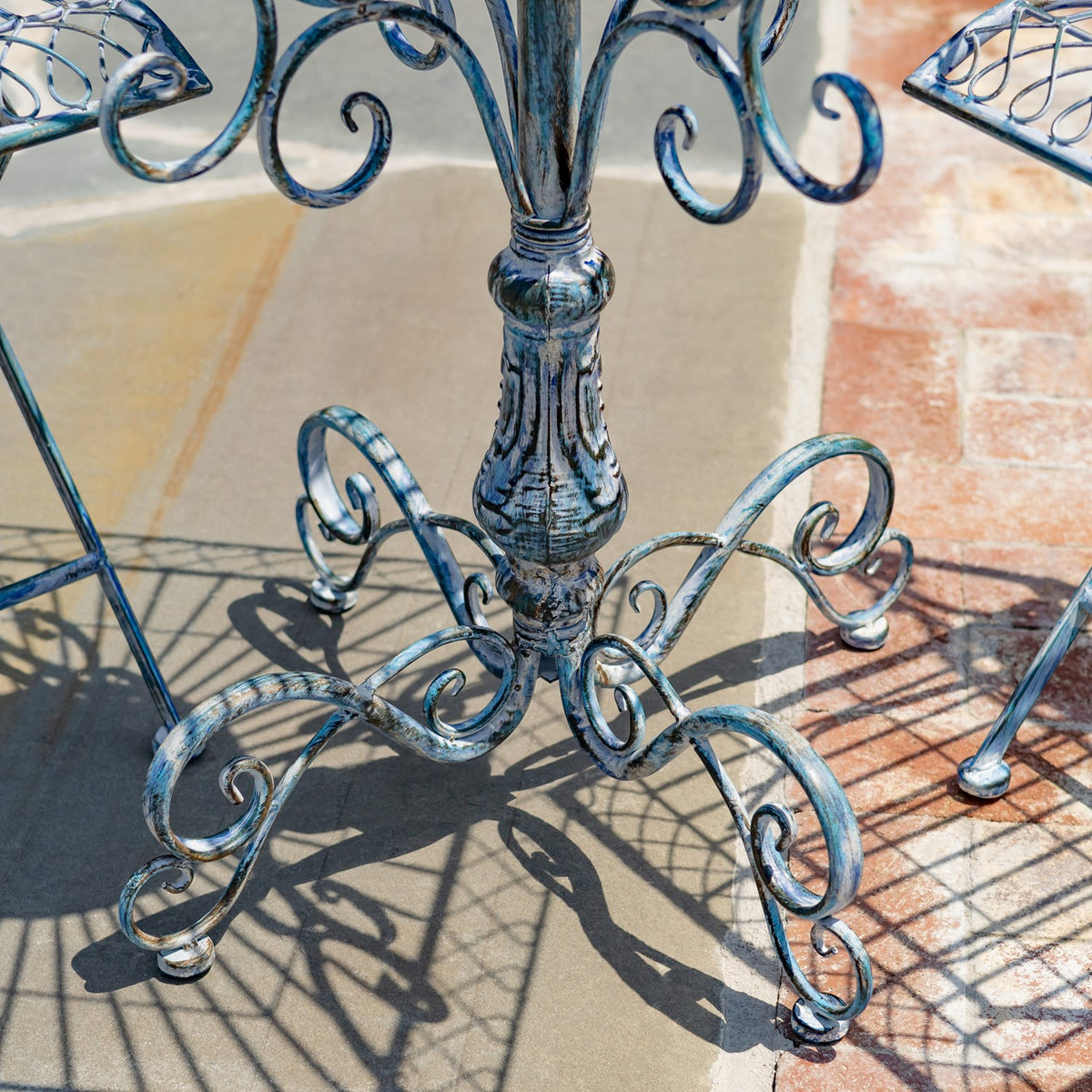Close-up of a distressed blue metal bistro table base with ornate curled feet and shadows on the patio.