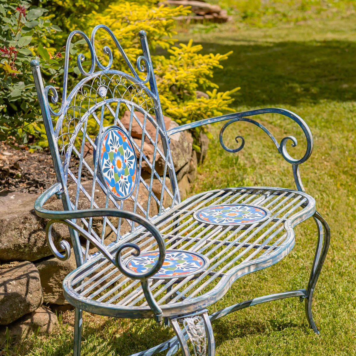 Ornate metal garden bench with colorful mosaic accents set on grass among shrubs and stones.