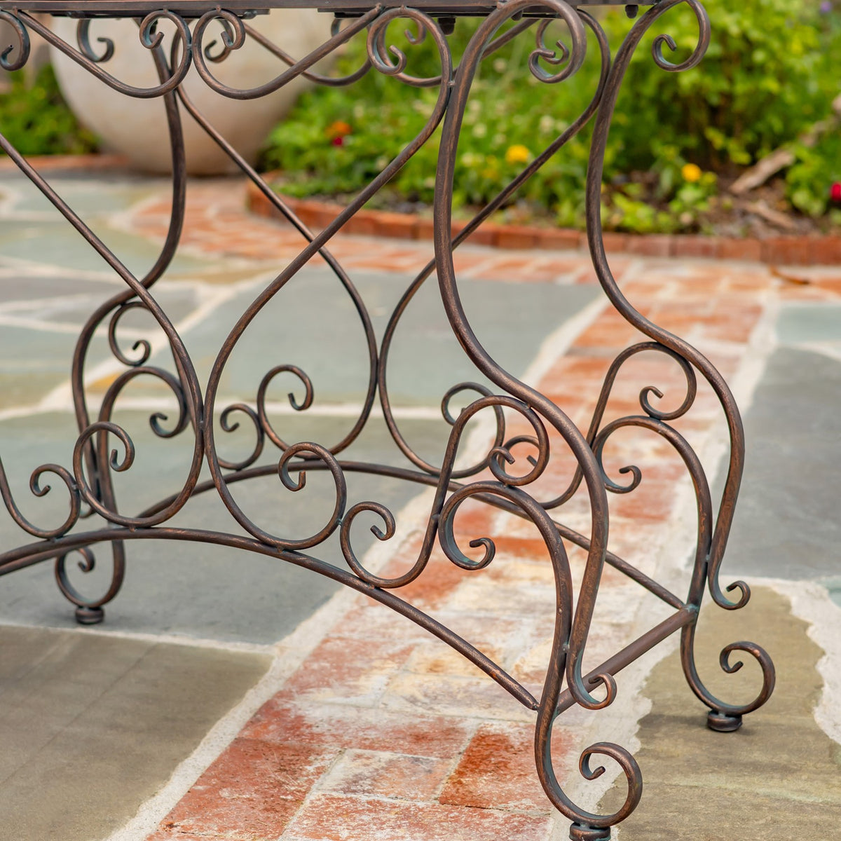 Close view of the plant stand’s curling bronze-colored metal legs and decorative scrollwork on a tiled patio.