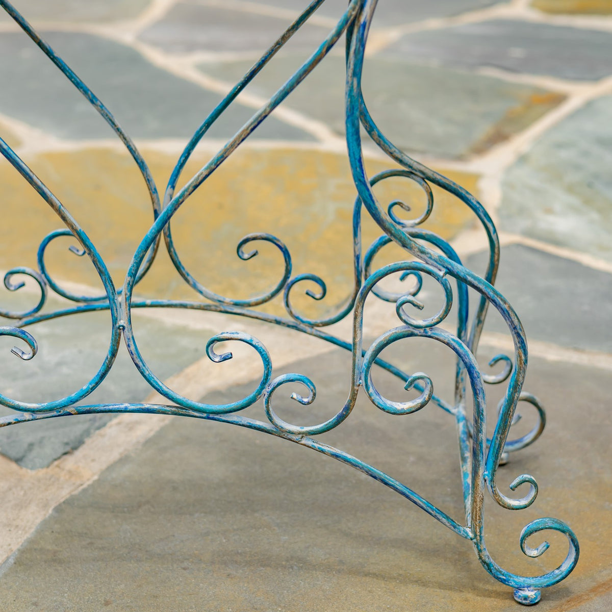 Curving blue metal scrollwork and slender legs of a plant stand against a stone-paved floor.