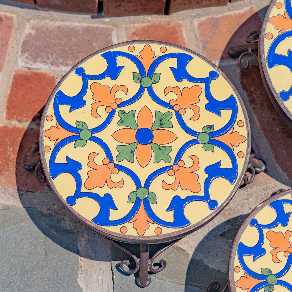 Round plant stand with a colorful floral mosaic top and curled metal legs, beside partial views of matching stands.