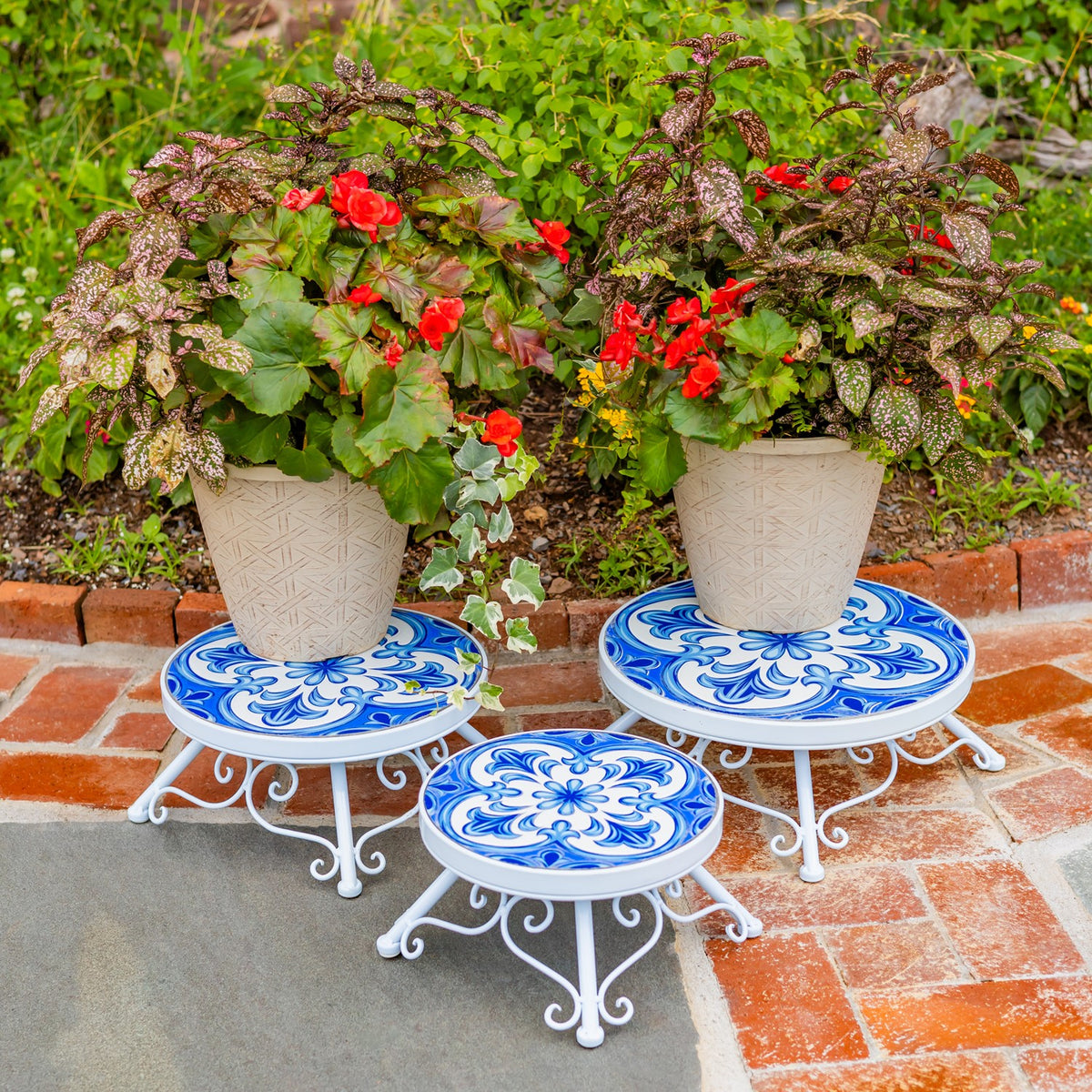 Three white metal plant stands with blue floral mosaic tops hold potted flowers on a brick patio.