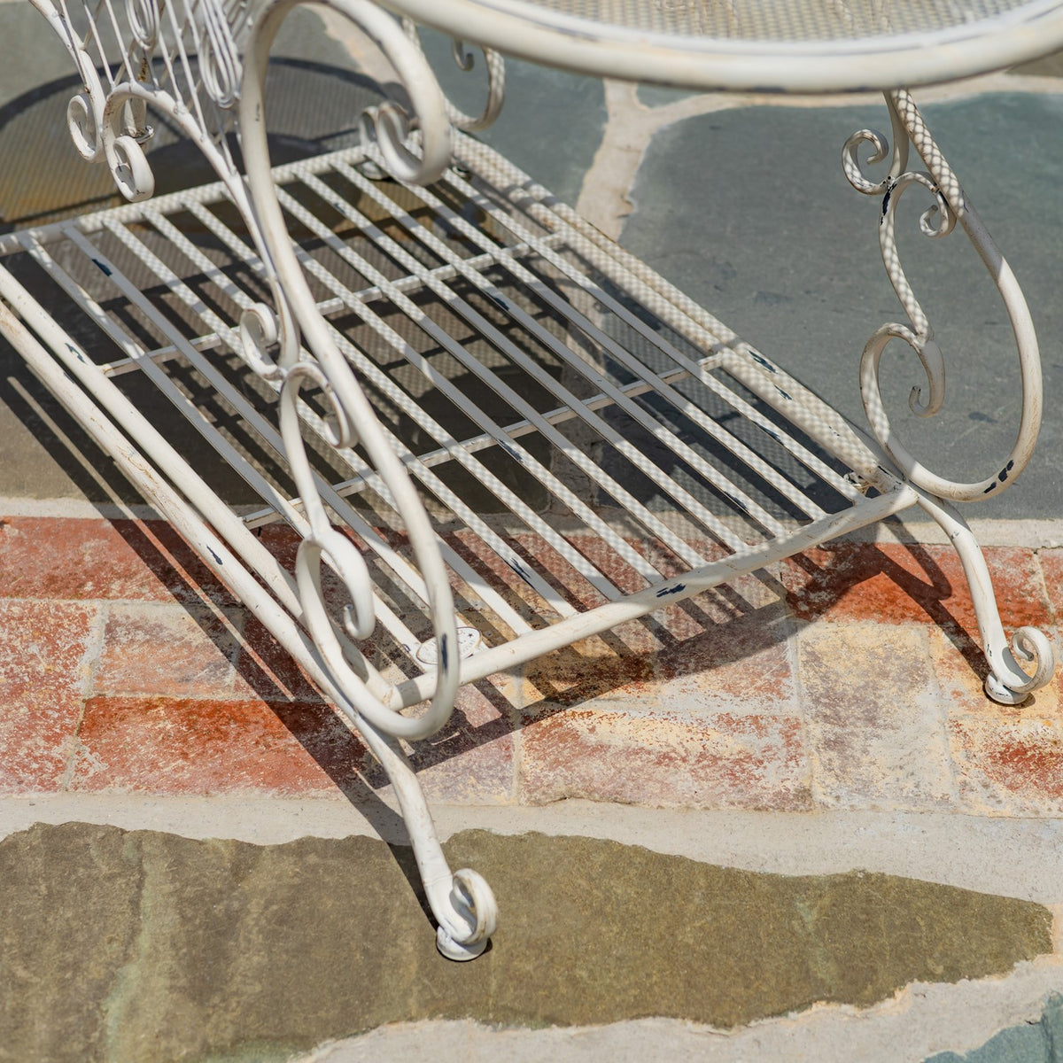 Close view of the table’s white metal lower slatted shelf and curled legs on a stone patio.
