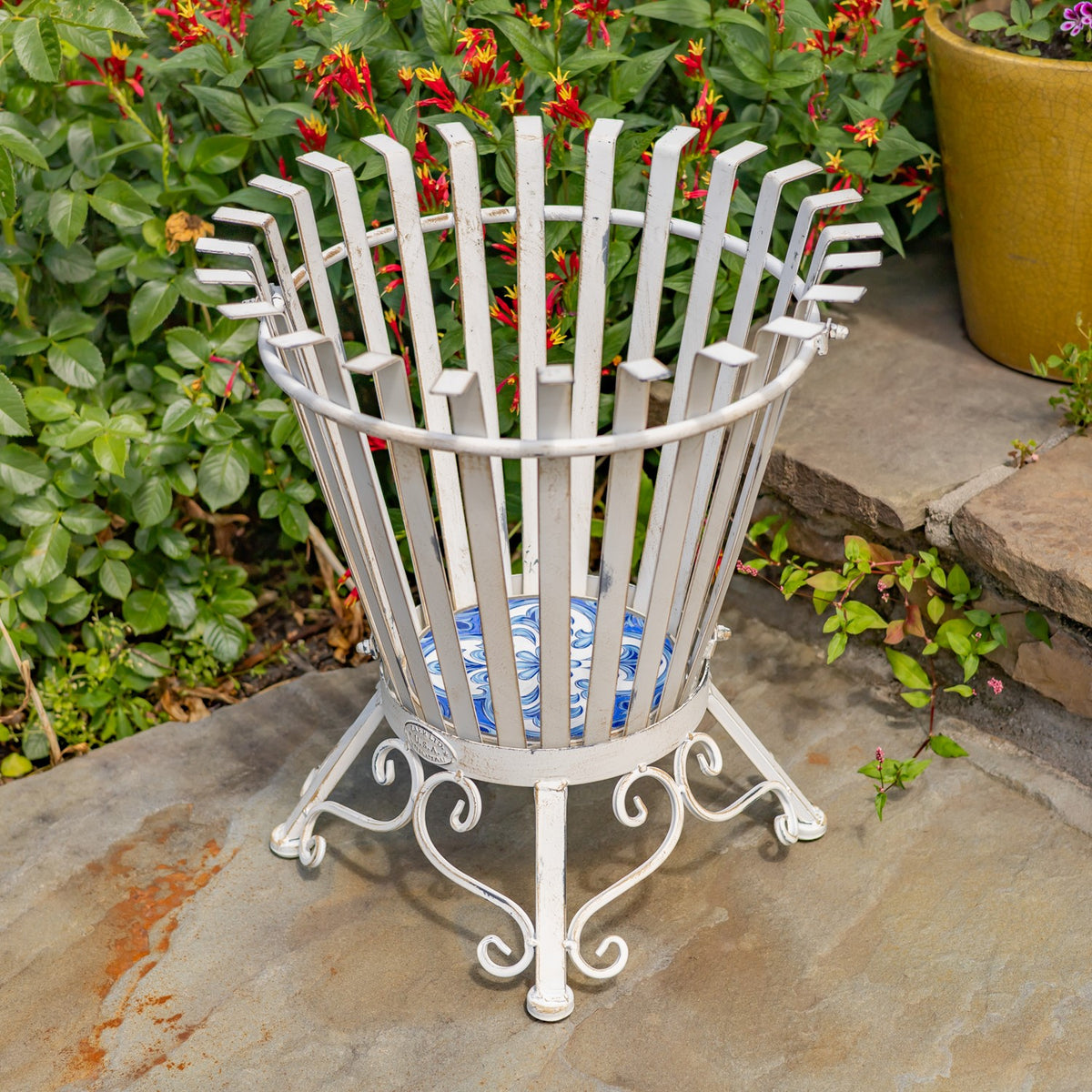 Antique-white metal firewood holder with a blue patterned mosaic base, shown outdoors among plants.