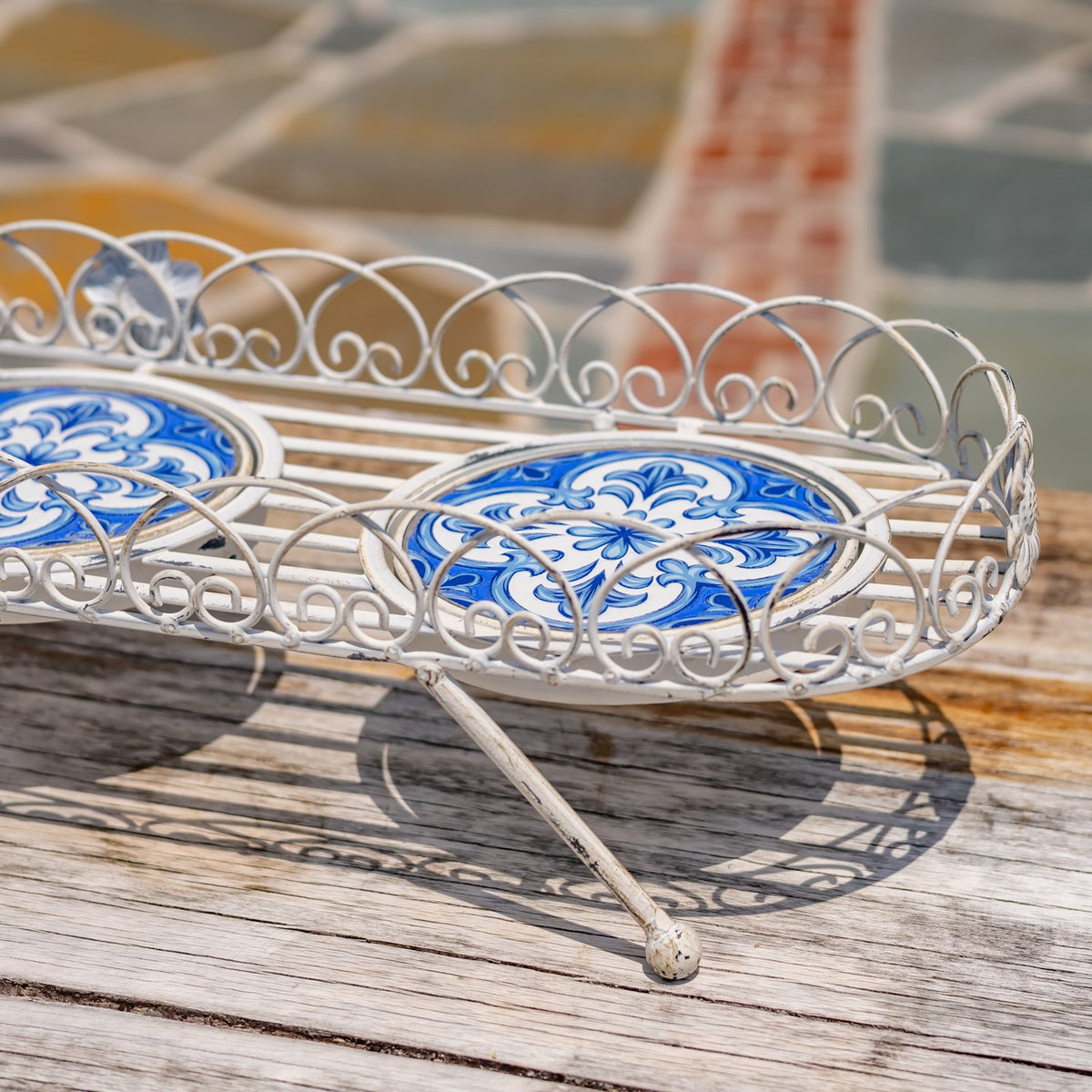 Antique-white scrollwork tray stand with blue-and-white mosaic insets on a wooden deck.