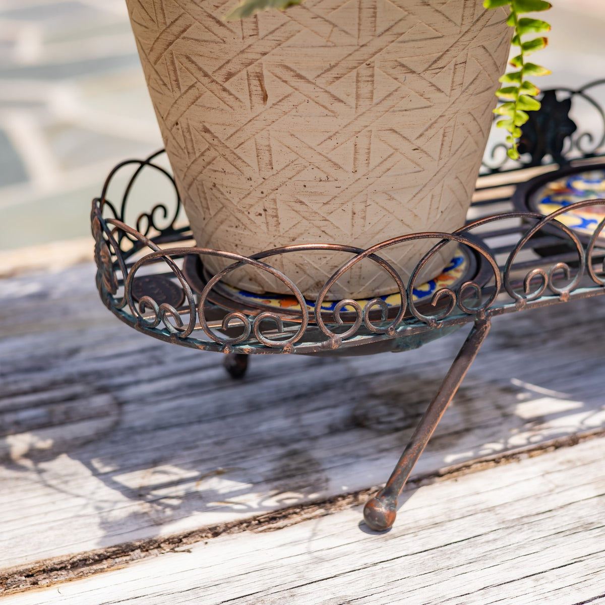Close-up of a patterned planter resting in a bronze-toned metal tray with scrollwork edging and a curved leg.