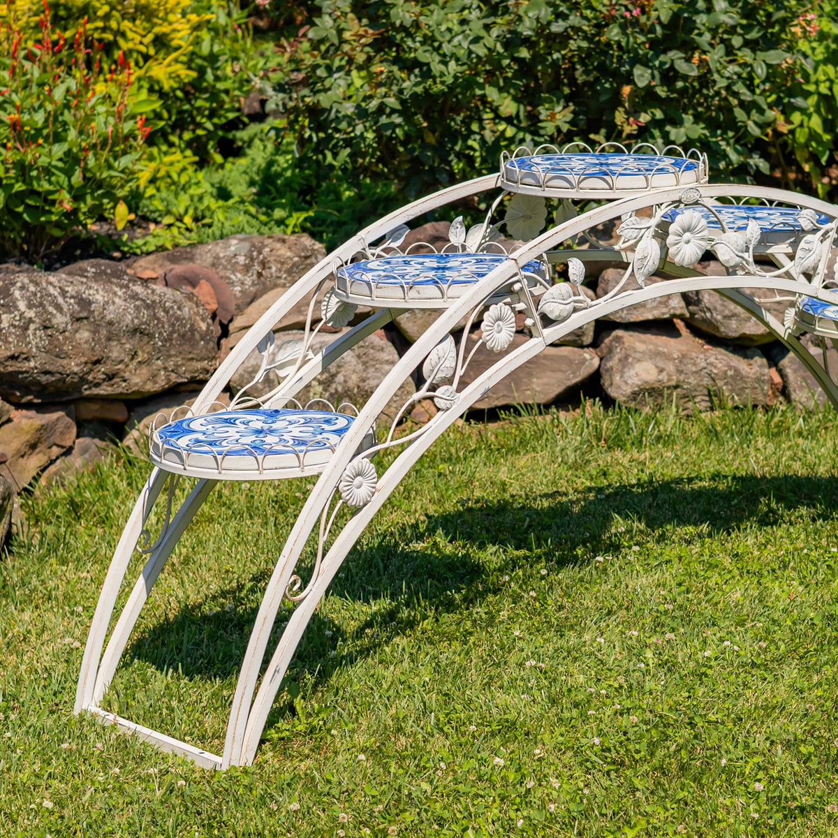 Arched white metal plant stand with blue patterned mosaic platforms set on a lawn beside a stone border.